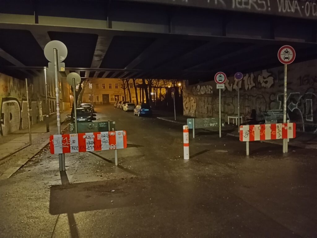 Auf dem Bild ist eine dunkle Straße zu sehen, die in der Mitte durch einen Poller und an den Seiten mit Rot-Weißen-Hindernissen bloockiert ist.